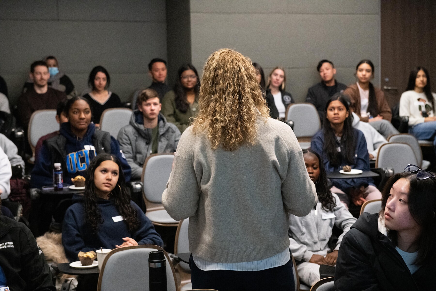 A person with long, blond hair stands face out on a diverse audience of teenagers