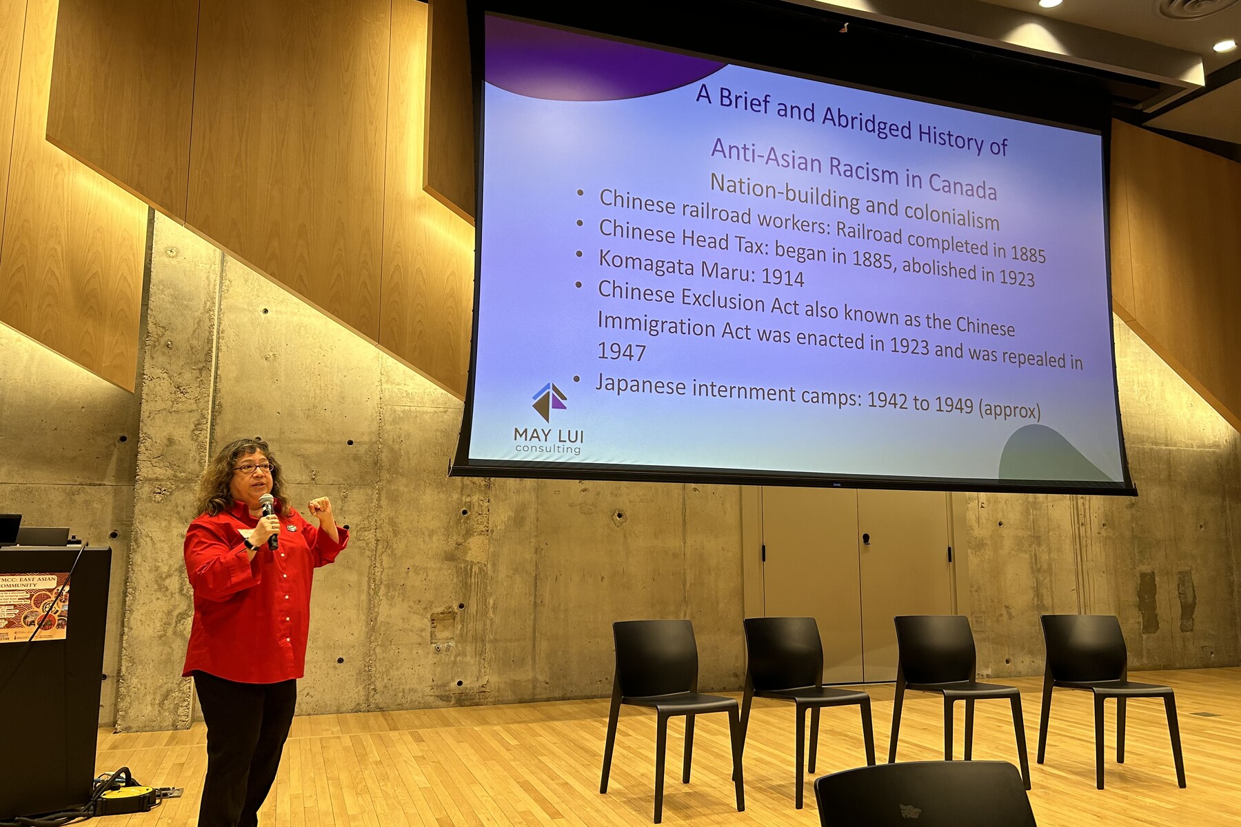 A woman stands at the front of a room with a microphone facing an audience, a screen and four chairs are to her left.