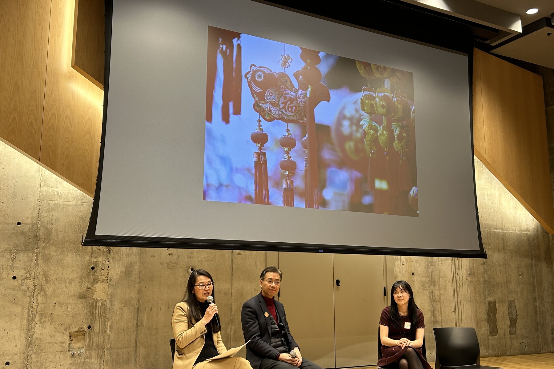 Three people sitting in chairs below a projector screen. The person on the person on the left holds a microphone.