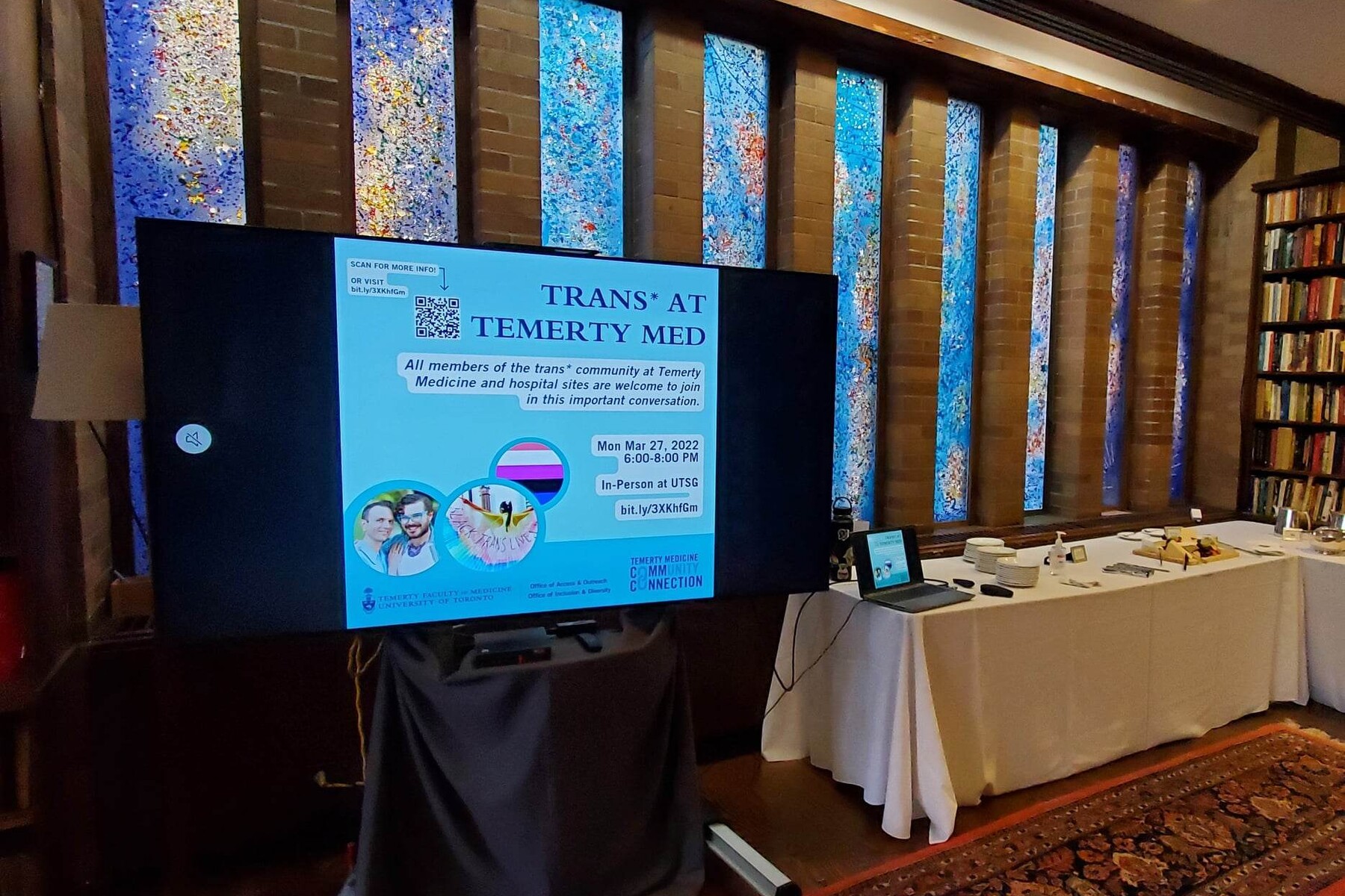 A poster on a large monitor displaying a promotion for the Trans* at Temerty event. To the right are tables with refreshments.