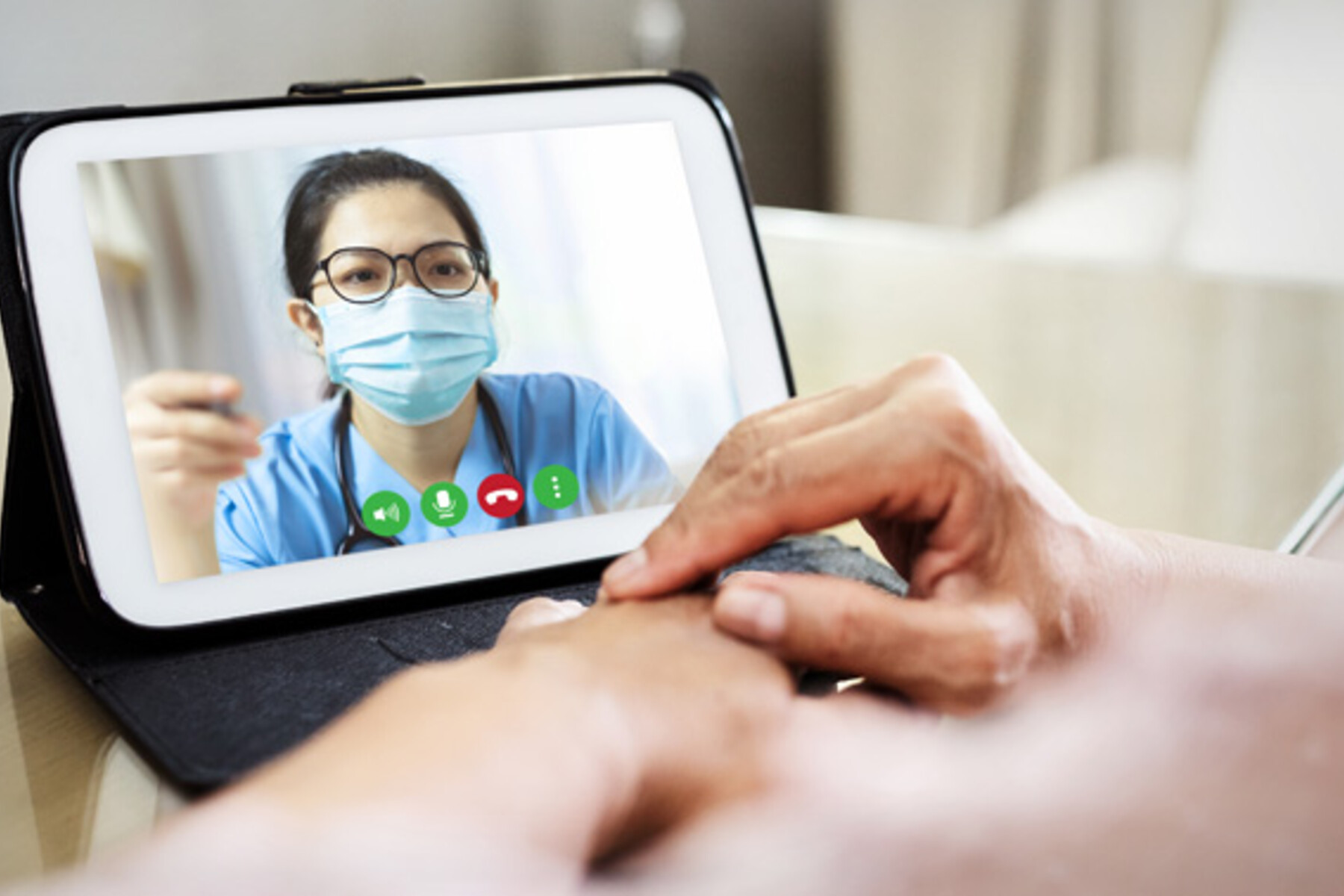A health care provider wearing a blue surgical mask is seen speaking to a another person via video chat on a tablet.