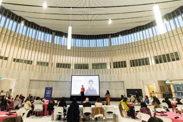 A room of people sitting at tables watching a speaker standing in front of a screen.