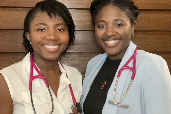 Ida (left) and Petra (right) stand side by side in front of a wood panelled background. They are wearing matching pink stethoscopes