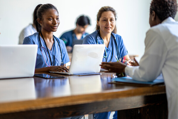 Two researchers in scrubs sit at a wooden table across from a doctor in a white coat.