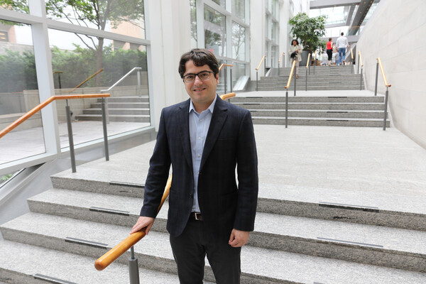 Daniel De Carvalho wearing a dark suit and a light blue shirt standing in a well-lit hallway with stairs
