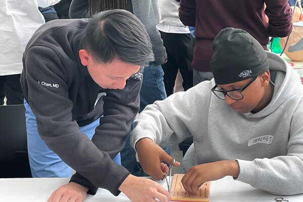 An adult in a dark grey sweater guides a teenager wearing glasses, a grey hoodie and beanie in a detailed activity at a table, with others present in the background.