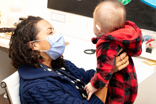 Suzanne Shoush, wearing a blue sweater and surgical mask, holds up a young baby dressed in a red plaid onesie.