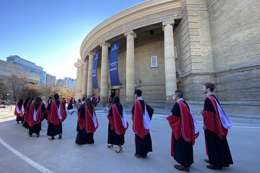 Graduands from the Temerty Faculty of Medicine wearing their gowns walk in a line outside Convocation Hall on a sunny fall day.