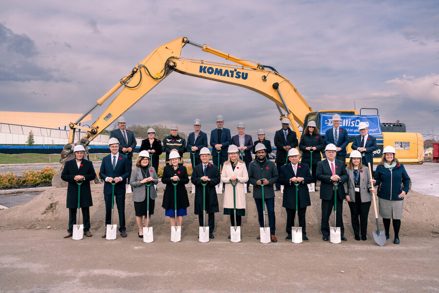 Stakeholders hold shovels and wear hard hats in front of a large yellow digger at SAMIH groundbreaking ceremony.
