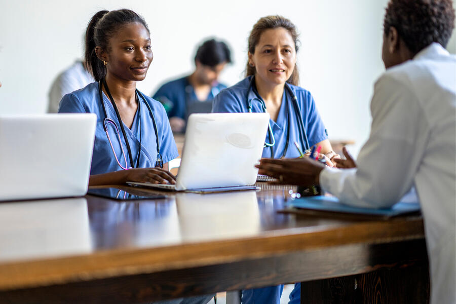 Two researchers in scrubs sit at a wooden table across from a doctor in a white coat.
