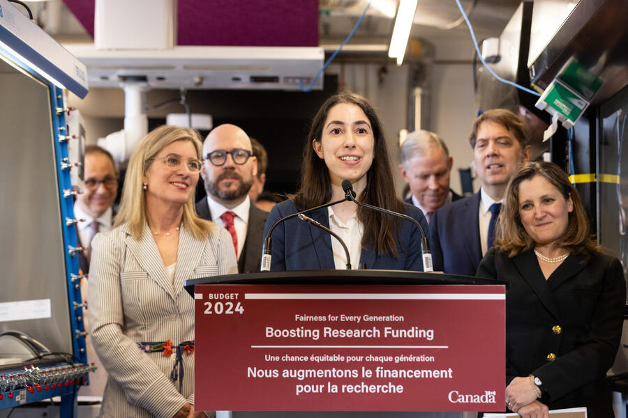 Kaitlin Kharas speaking at a podium with a sign that reads "Fairness for every generation. Boosting research funding."