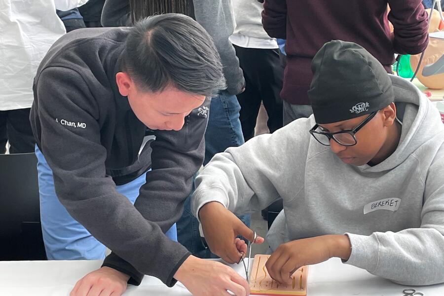 An adult in a dark grey sweater guides a teenager wearing glasses, a grey hoodie and beanie in a detailed activity at a table, with others present in the background.