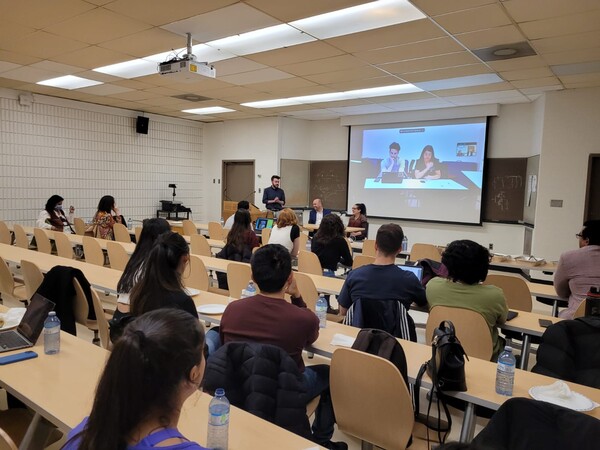 A photo of students in a classroom, with members of the HMSA presenting at the front in-person or and on-screen.