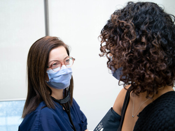 Professor Danielle Martin speaks to a patient and checks their blood pressure. Both wear blue surgical masks. Martin wears glasses.