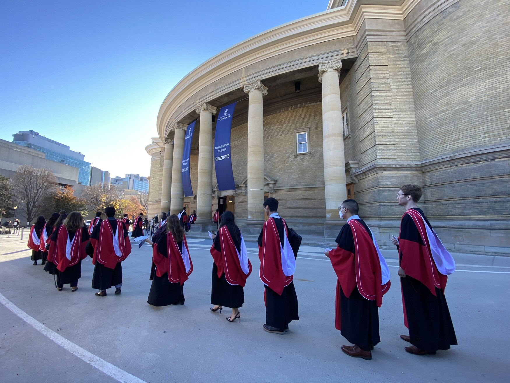 Graduands from the Temerty Faculty of Medicine wearing their gowns walk in a line outside Convocation Hall on a sunny fall day.