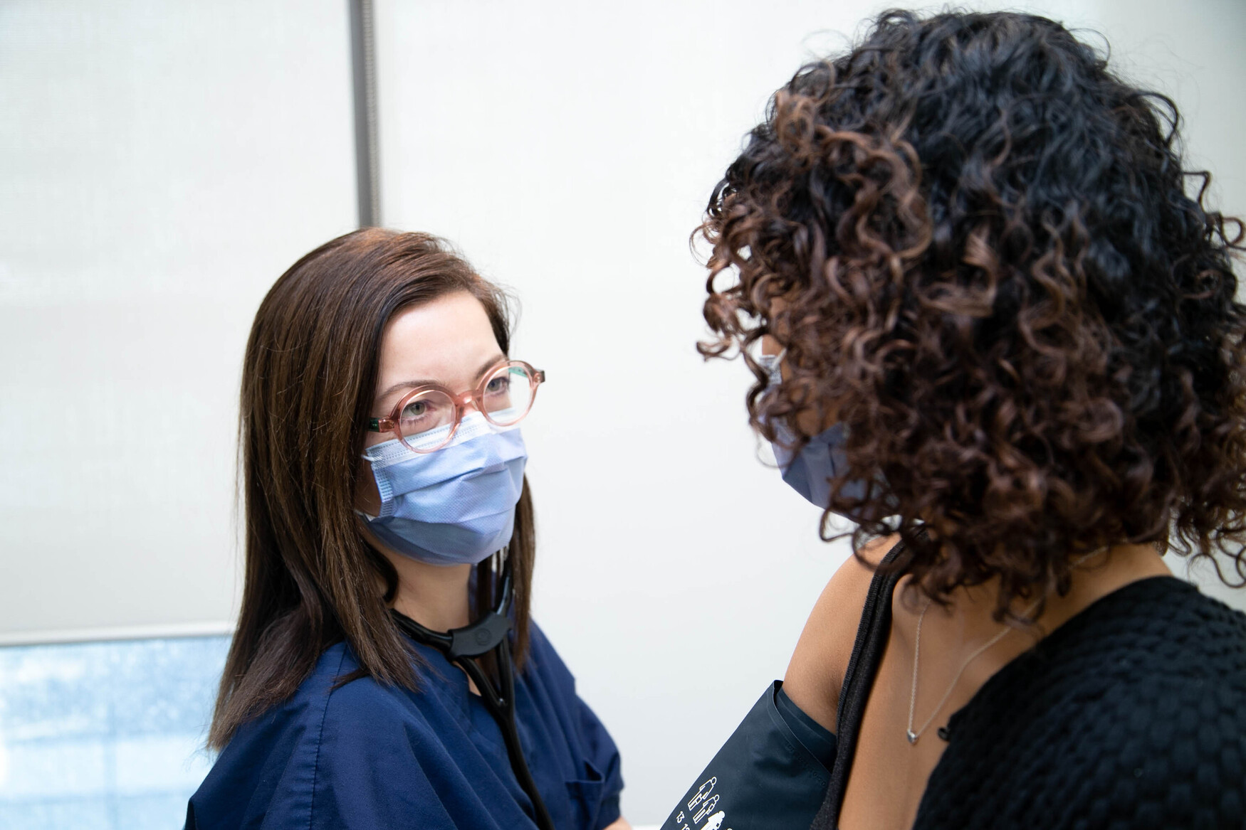 Professor Danielle Martin speaks to a patient while checking their blood pressure. Both wear blue surgical masks. Marin also wears glasses.