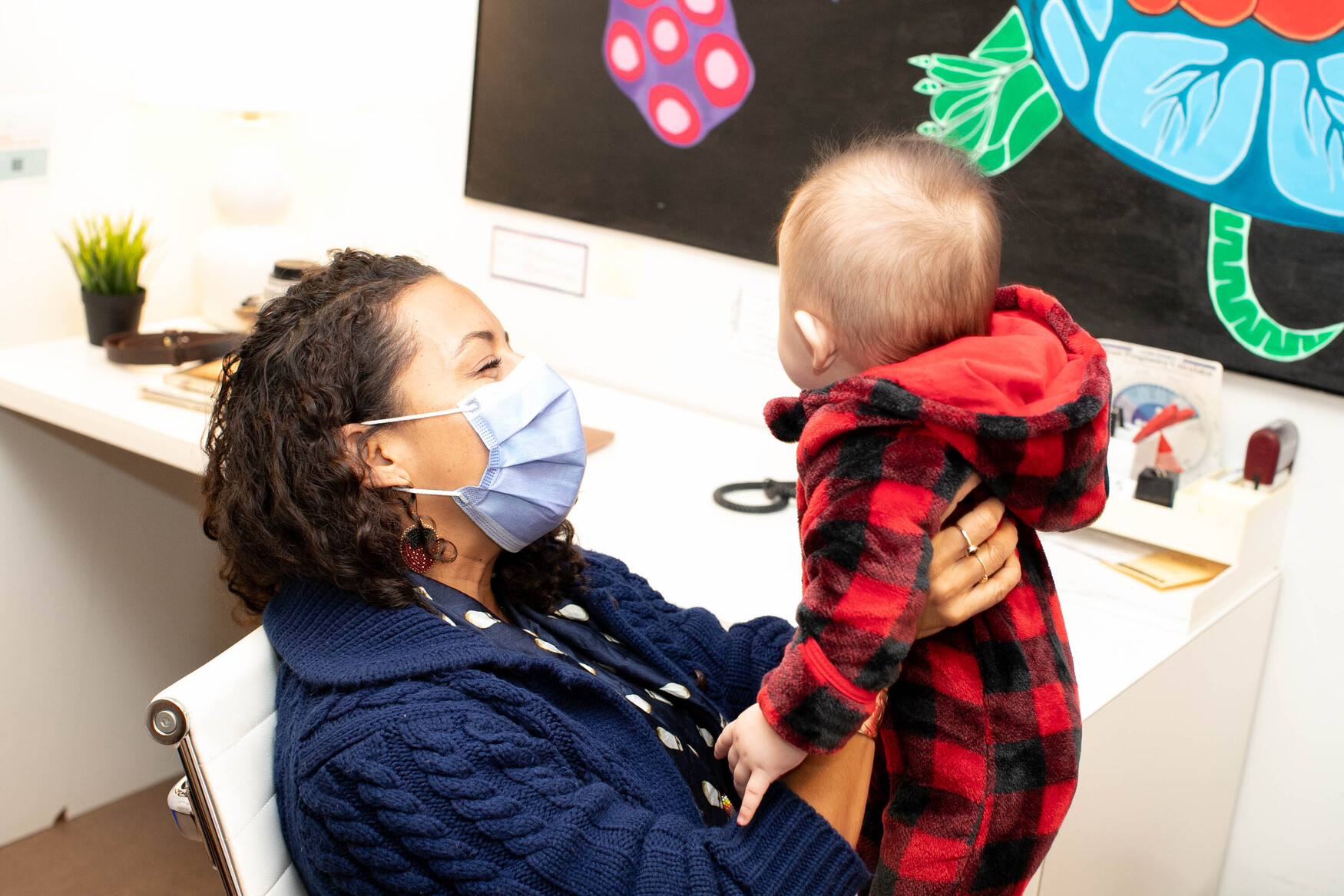 Suzanne Shoush, wearing a blue sweater and surgical mask, holds up a young baby dressed in a red plaid onesie.