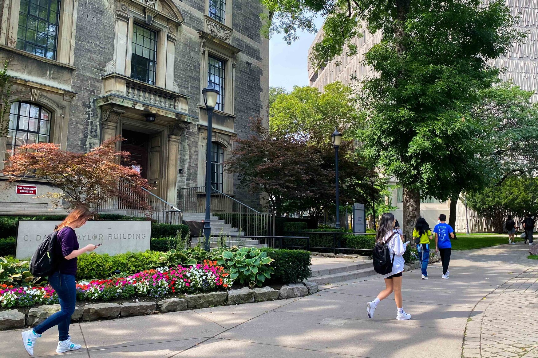 People walk on the path in front of the Naylor Building on the U of T St. George Campus on a sunny, warm day. 