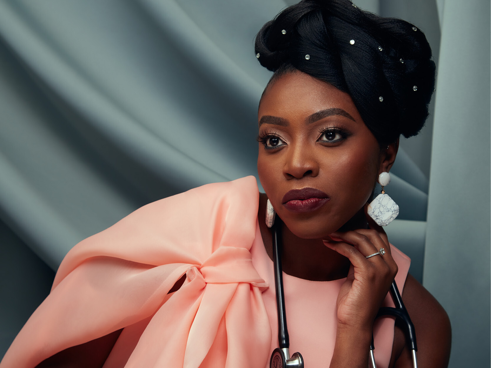 Chika Oriuwa, MD, poses in front of a grey back drop. She is wearing a pink blouse with a one large rouched sleeve on her right shoulder and has a black stethoscope around her neck 