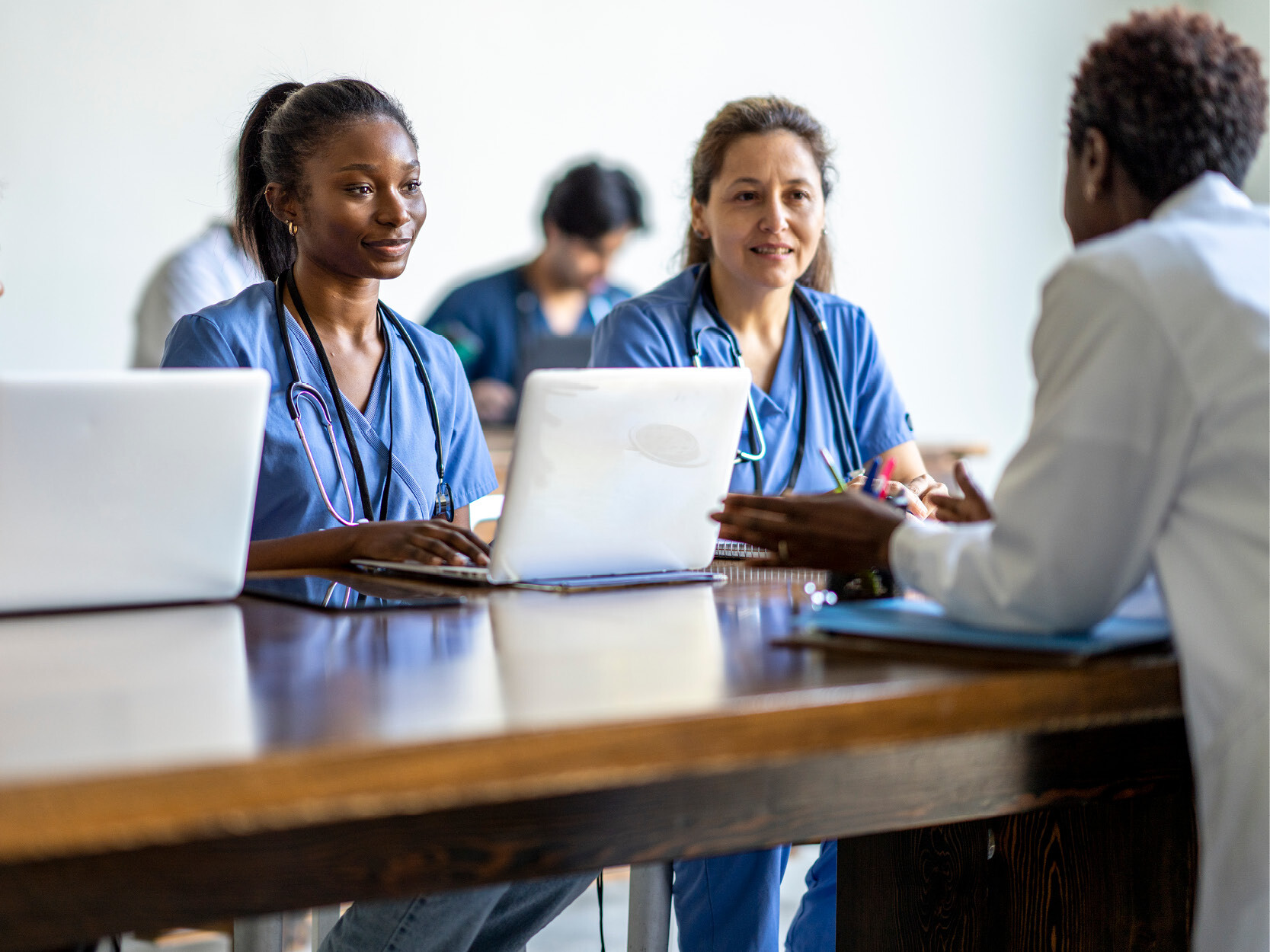 Two researchers in scrubs sit at a wooden table across from a doctor in a white coat.