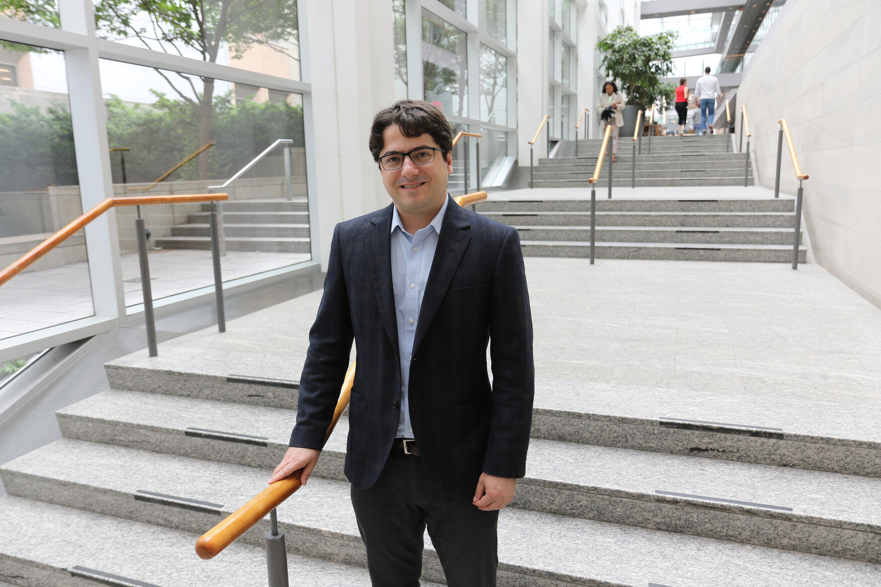 Daniel De Carvalho wearing a dark suit and a light blue shirt standing in a well-lit hallway with stairs
