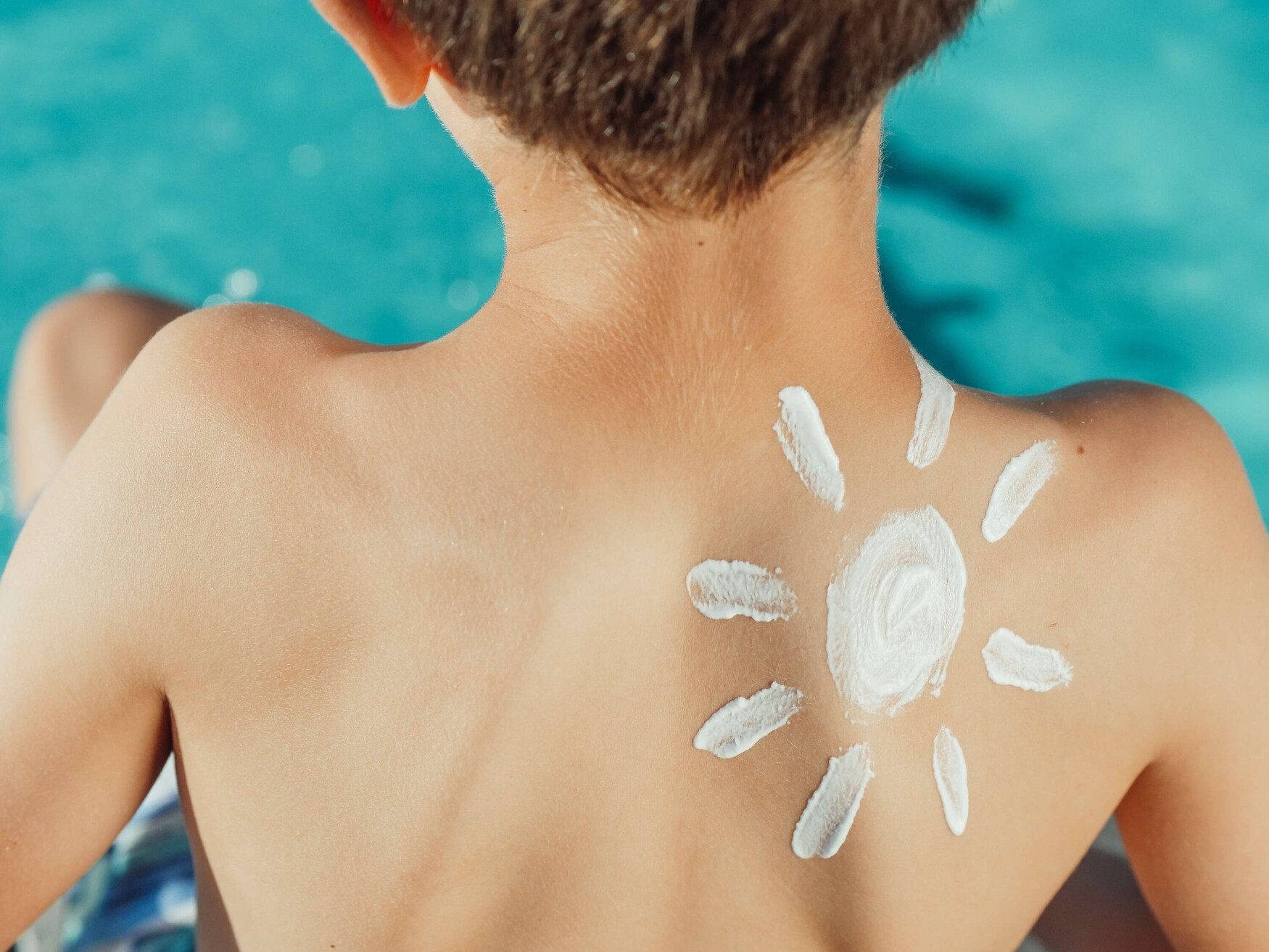 Photo of the back of a light skinned boy beside a pool with a sun sketched on his shoulder in white sunscreen. Photo by Kindel Media from Pexels