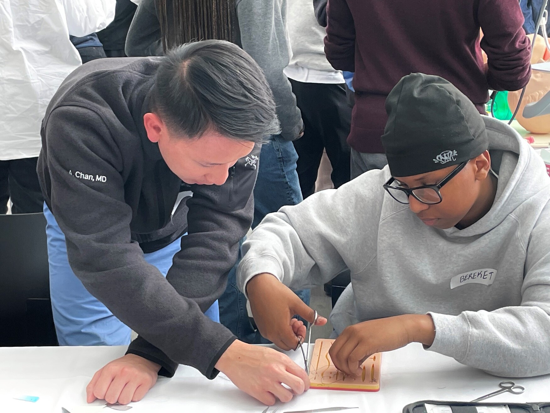 An adult in a dark grey sweater guides a teenager wearing glasses, a grey hoodie and beanie in a detailed activity at a table, with others present in the background.