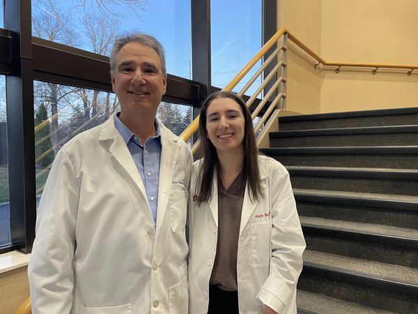 a man and a young woman both wearing lab coats smile into the camera