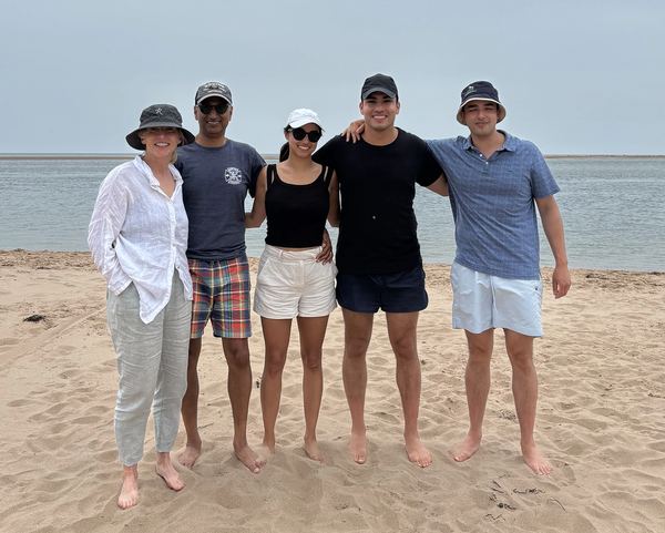 A family of five adults stand on a beach and smile into the camera