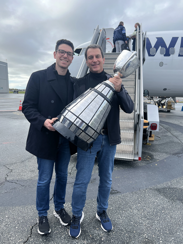 two men hold the Grey Cup trophy and smile into the camera